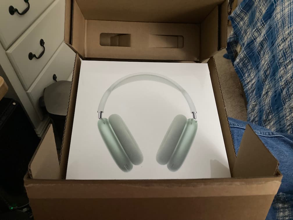 the two folded covers pulled back, revealing the shrink wrap sealed AirPods Max package, in green color